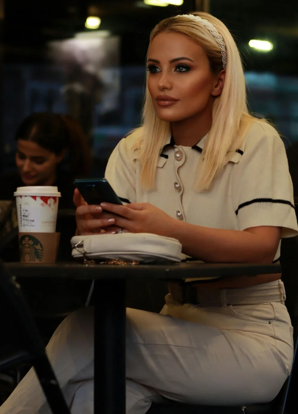 Une jeune femme est assise à une table de café, souriant en regardant son téléphone. Un homme s'approche d'elle et ils engagent la conversation.